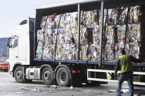 A large commercial waste truck operated by Commercial Waste Removal Erith is parked on a paved area, fully loaded with compressed, mixed waste materials such as paper, cardboard, plastic, and other household or office refuse, visibly tightly packed within an open-sided, steel-framed container. The waste appears in various colours and textures, with crumpled and flattened forms and some loose debris at the truck's rear, indicating recent loading. To the right of the truck, a person wearing a high-visibility vest and casual clothing is walking past, holding a small object or mobile device. The background features a clear, bright sky with natural daylight illuminating the scene. In the distance to the left, part of a vehicle and other infrastructure elements are visible, suggesting a commercial or industrial area, possibly near Erith or Kent postcode region. This scene exemplifies the type of rubbish collection services offered by Commercial Waste Removal Erith, focusing on efficiently managing bulky waste loads and debris collection in the local area.