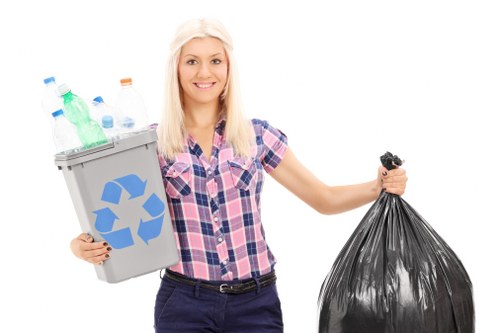 A young woman with long blonde hair, wearing a pink and blue checkered shirt and dark trousers, is smiling and standing against a plain white background. She is holding a grey recycling bin filled with plastic bottles of various shapes and colors, including green, blue, and clear, with the recycling symbol displayed on the front. In her other hand, she is gripping a tightly tied black plastic rubbish bag. The image focuses on her, the recycling bin, and the rubbish bag, representing waste disposal and rubbish removal services. The scene conveys a neat and organized approach to waste management, suitable for a professional rubbish removal company operating in a residential or commercial context, such as Erith or nearby areas in London. The clean, well-lit setting emphasizes environmental responsibility and proper waste segregation, aligning with the services offered by Commercial Waste Removal Erith.