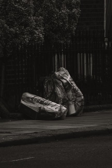 A black-and-white photograph showing several large plastic rubbish bags placed on the pavement next to a black metal fence, with dense foliage of a tree visible behind the fence. The bags appear to co