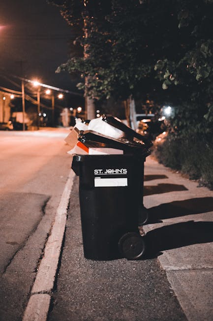 A black-and-white photograph showing several large plastic rubbish bags placed on the pavement next to a black metal fence, with dense foliage of a tree visible behind the fence. The bags appear to co