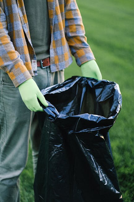 Two workers dressed in high-visibility orange and red safety clothing are performing rubbish removal on a busy city street. They are pushing a wheeled bin full of waste towards a collection point. The