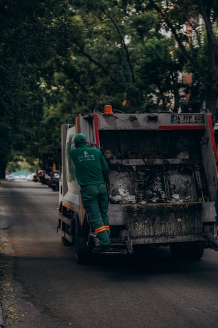 A worker dressed in a green uniform with orange reflective stripes on the pants is standing next to an open back of a small rubbish collection van on a suburban street. The van, painted white with som