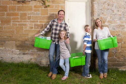 A family of four standing outdoors on a grassy area in front of a brick and stone wall, each holding a bright green plastic bin used for rubbish collection or waste disposal. The father, dressed casually in a plaid shirt and jeans, stands on the left with a smile and a bin in each hand; a young girl with curly hair, wearing a grey sweater and jeans, stands in front of him, also holding a bin. To the right, a boy dressed in a blue and white striped shirt and dark trousers holds a bin, and a woman wearing a white blouse and jeans stands on the far right, holding another green bin. The scene is well-lit, with natural daylight highlighting the textured brick and stone background, which suggests a residential setting, possibly in Erith, relevant to local rubbish removal services. The placement of the family emphasizes the community aspect of rubbish collection and waste management facilitated by Commercial Waste Removal Erith, with visual cues pointing to waste disposal at a domestic or small commercial property.