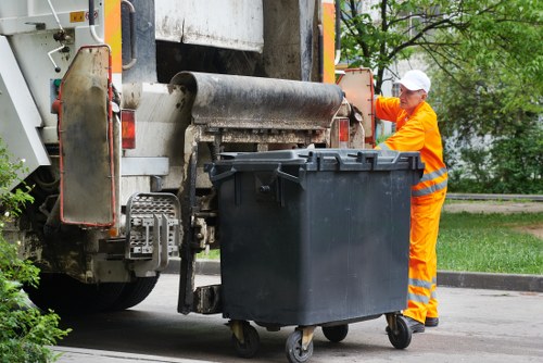 A worker dressed in a high-visibility orange and yellow safety uniform, including a white cap, is operating a large waste collection vehicle on a paved street in Erith. The worker is positioned beside a black wheeled rubbish bin, which is partially in contact with the vehicle's loading mechanism. The vehicle, primarily grey with orange safety accents, appears to be a commercial waste collection truck used for rubbish removal services. The scene is set outdoors, with greenery and trees visible in the background, indicating it is a daytime operation in a suburban or urban area. The truck's lifting and loading equipment is engaged in emptying the bin, highlighting a routine rubbish collection process. The environment suggests an organized waste disposal activity, typical for commercial rubbish removal services in the Erith area, supported by a professional attendant ensuring safe handling of waste. The overall atmosphere is one of efficiency and routine waste management activity, with a focus on proper waste collection logistics.