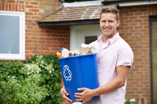 A young man with short light brown hair, wearing a light pink short-sleeved shirt, is standing outdoors in front of a brick house with a white window frame and a tiled roof. He is smiling and holding a blue recycling bin made of plastic, which is filled with various types of rubbish including paper, cardboard, and plastic bottles. The bin features a white recycling symbol on its side. In the background, there is a green shrub with white flowers and a paved driveway or garden area. The environment is bright and sunny, suggesting a residential setting suitable for rubbish collection and waste removal services offered by Commercial Waste Removal Erith near the postcode district of Erith.