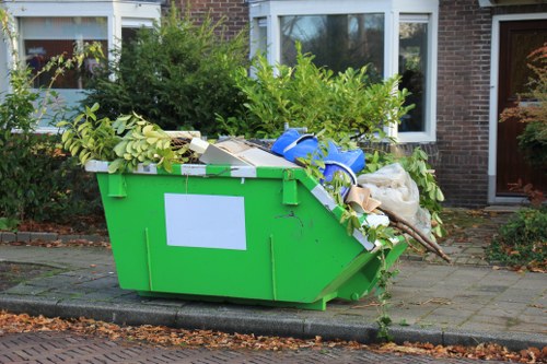 A large, bright green skip bin positioned on a paved driveway outside a residential property, filled with a mix of garden waste such as leafy branches, green foliage, and twigs, along with white packaging materials, cardboard boxes, and plastic items including a blue strap. The skip is situated on the edge of a concrete curb, with paving slabs extending to the house entrance, which features large windows and a brick exterior. Surrounding bushes and trees with green leaves are visible in the background, indicating a suburban environment. The scene appears to be daytime with natural light illuminating the area, emphasizing the textures of the waste materials and the skip's glossy finish. This setup is typical of rubbish removal services by Commercial Waste Removal Erith, serving properties in or near the local postcode area, emphasizing the collection of garden and domestic waste in an urban setting.